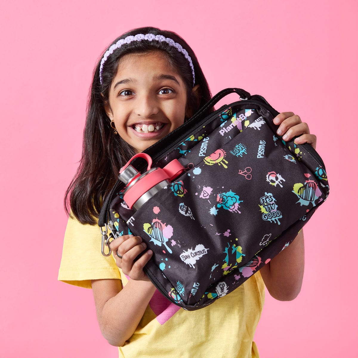 A young girl with dark hair and a bright smile holds up a PlanetBox Adventurer Insulated Lunch Bag in the Street Art Pattern - a colorful graffiti-inspired design against a pink background. The black lunch bag features vibrant doodles and positive messages. A stainless steel Glacier Chug Lid water bottle with a pink lid is tucked into the side pocket. She is wearing a yellow t-shirt and a white headband, radiating excitement.