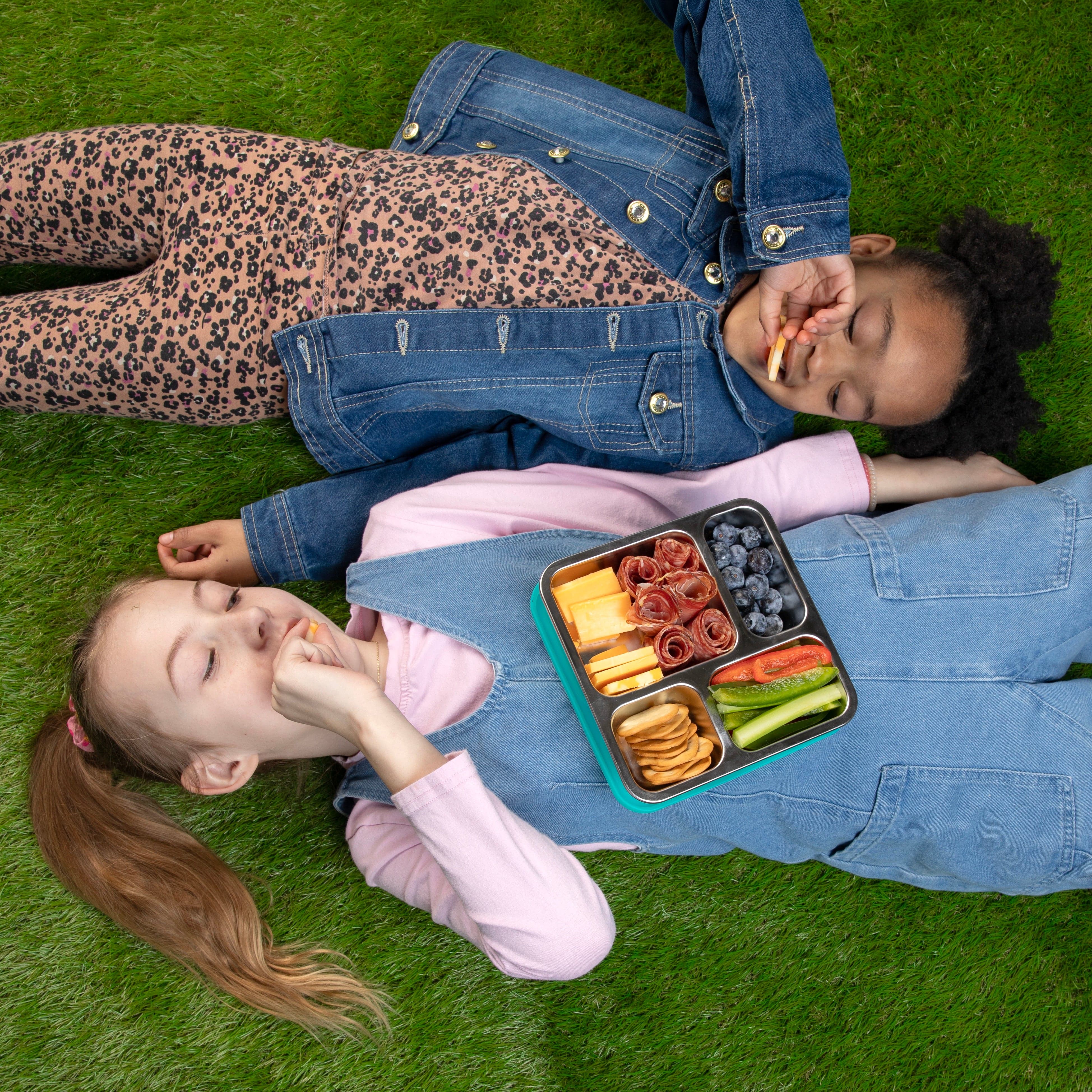 Two children lying on grass with a voyager lunch box containing snacks.
