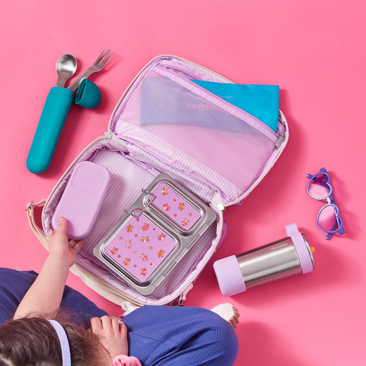 An open Adventurer Insulated lunch bag showing the interior organization and included items: stainless steel utensils in teal case, Pansy Day Tripper Snack Container, Stainless Steel Shuttle two compartment lunch box with Cats Meow magnets on top, a Coldkit ice pack in a mesh pocket, a stainless steel Glacier Flip Straw Water Bottle in Pansy, and purple heart sunglasses arranged around it, all arranged on a bright pink background. A child wearing blue clothing is partially visible in the bottom left corner.