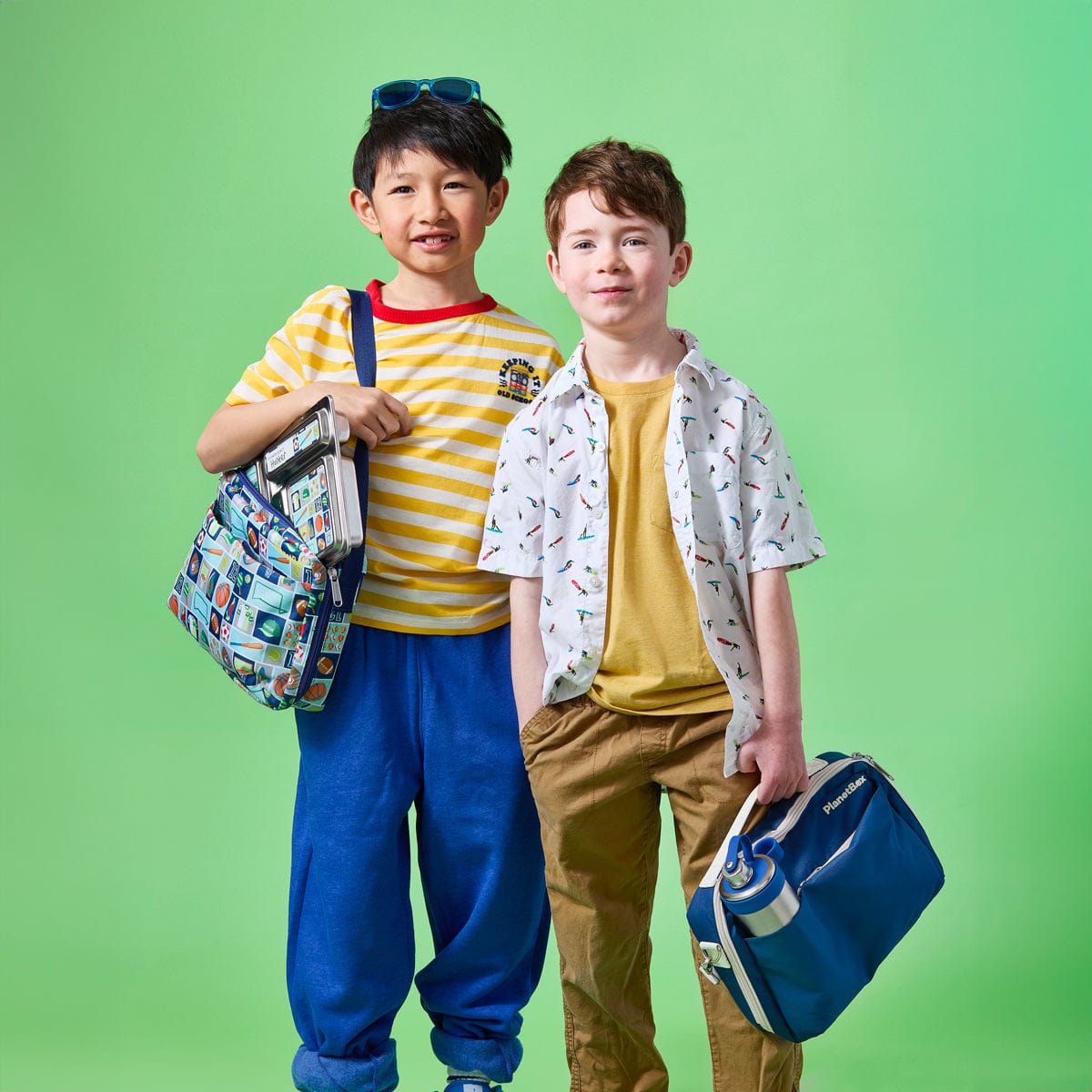 Two young boys, one with a patterned blue Adventurer Insulated lunch bag and the other with a plain blue Adventurer Insulated lunch bag, pose against a vibrant green backdrop. The boy on the left, who may be of East Asian ethnicity, wears a yellow striped shirt and blue pants. The boy on the right, who may be of Caucasian ethnicity, wears a white patterned shirt and brown pants.