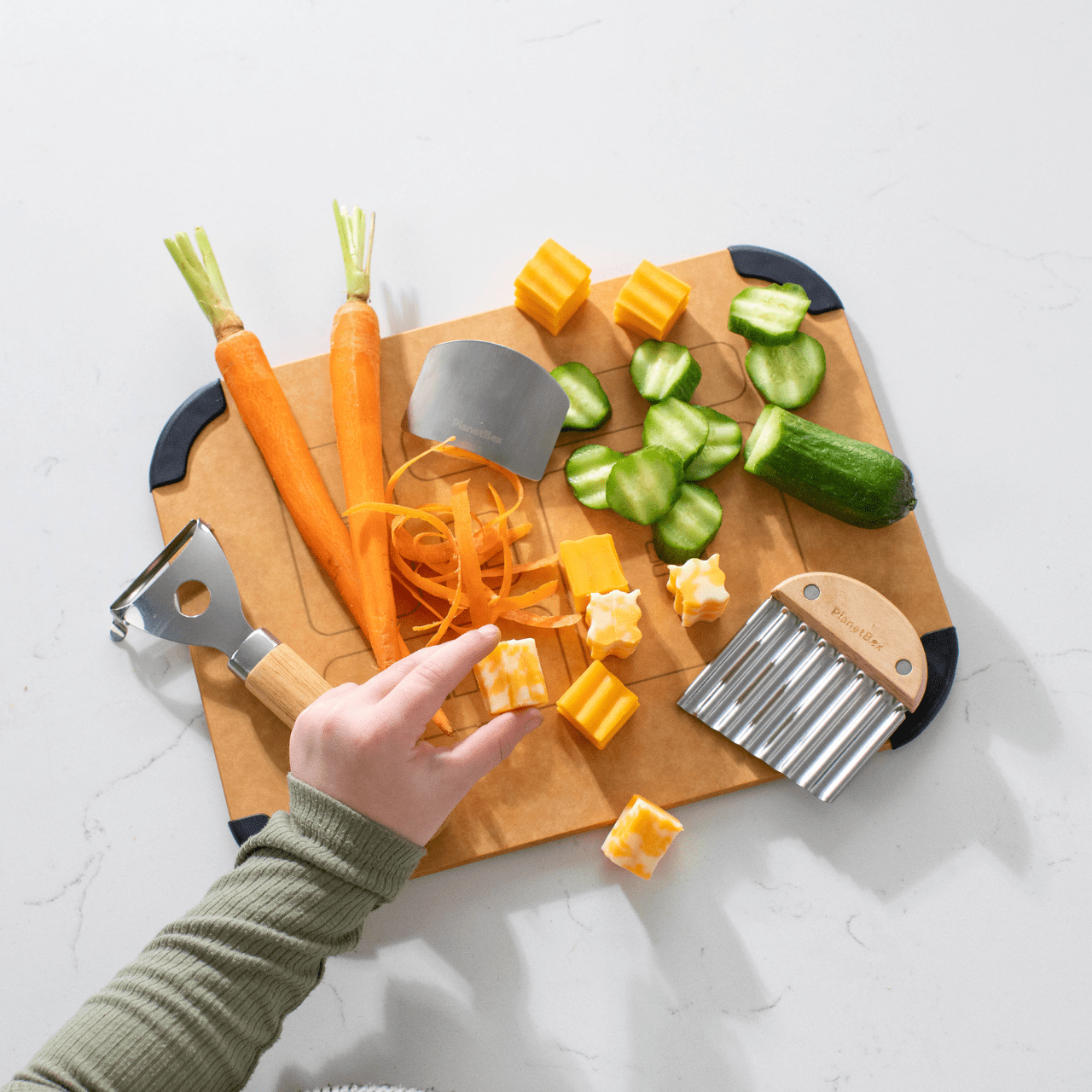 Prep to Pack Three Piece set laid on top of Prep to Pack cutting board with chopped cucumber, cheese and carrots. Childs hand is lifting cheese cube from the board.