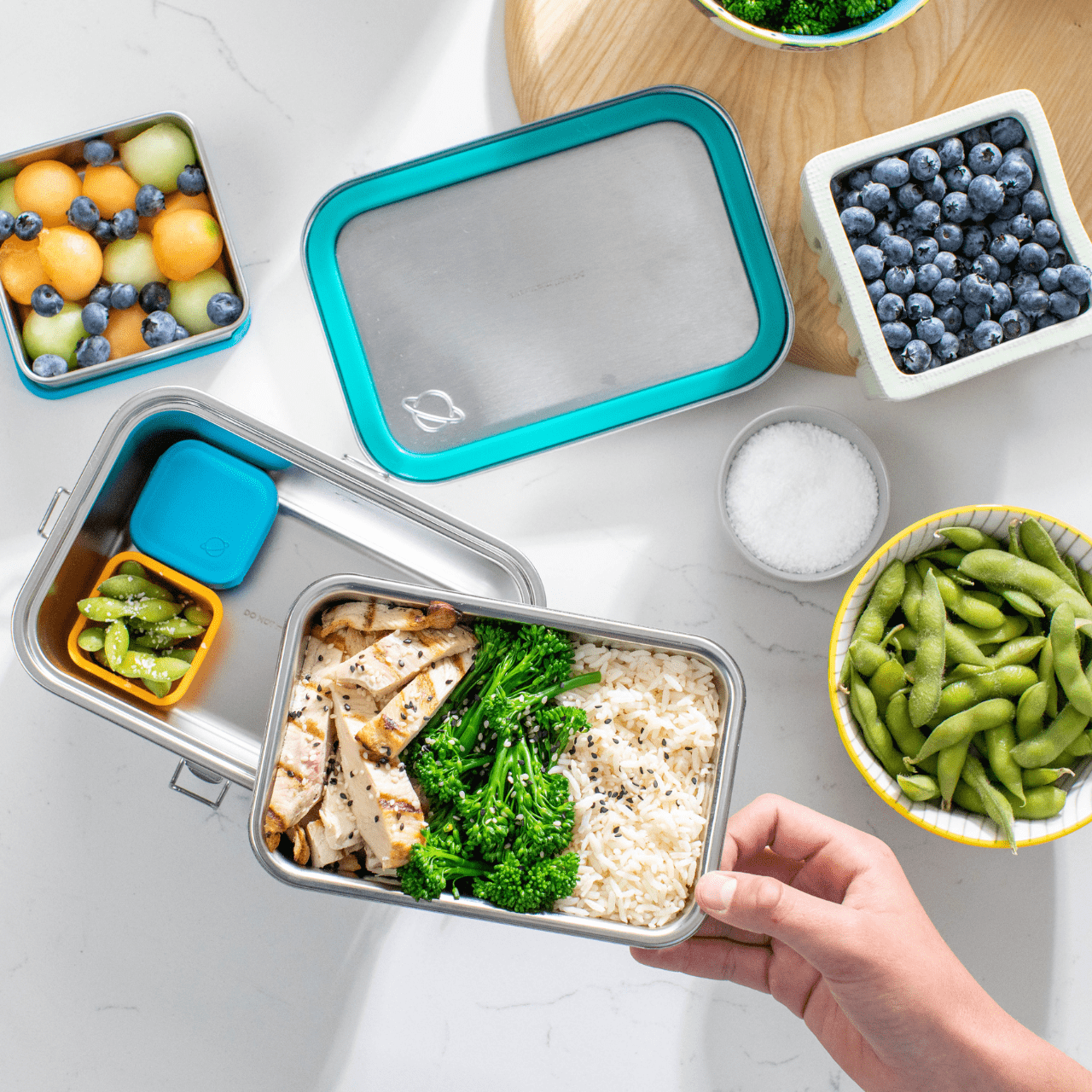 Disassembled Explorer lunchbox with dippers containing fruit, and pods containing edamame. Microwaveable tray includes broccoli, rice and chicken. Assorted fruits and vegetables in background.