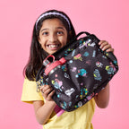 A young girl with dark hair and a bright smile holds up a PlanetBox Adventurer Insulated Lunch Bag in the Street Art Pattern - a colorful graffiti-inspired design against a pink background. The black lunch bag features vibrant doodles and positive messages. A stainless steel Glacier Chug Lid water bottle with a pink lid is tucked into the side pocket. She is wearing a yellow t-shirt and a white headband, radiating excitement.