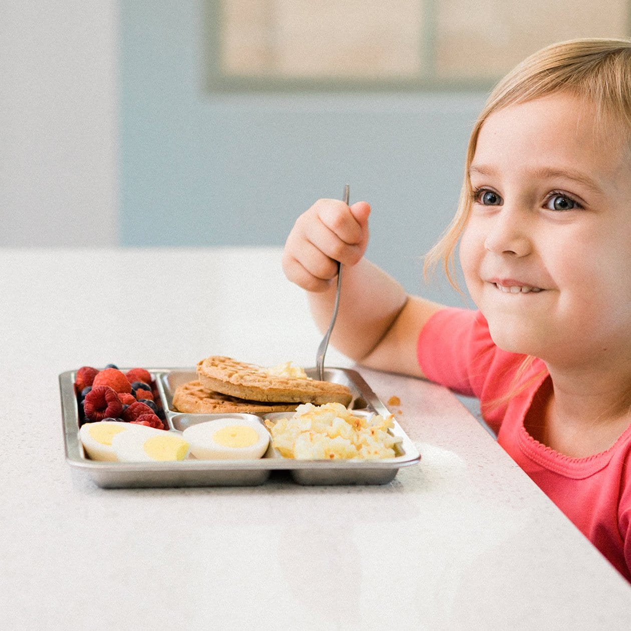 Child eating breakfast of waffles, berries, hardboiled egg and potatoes on tray. 