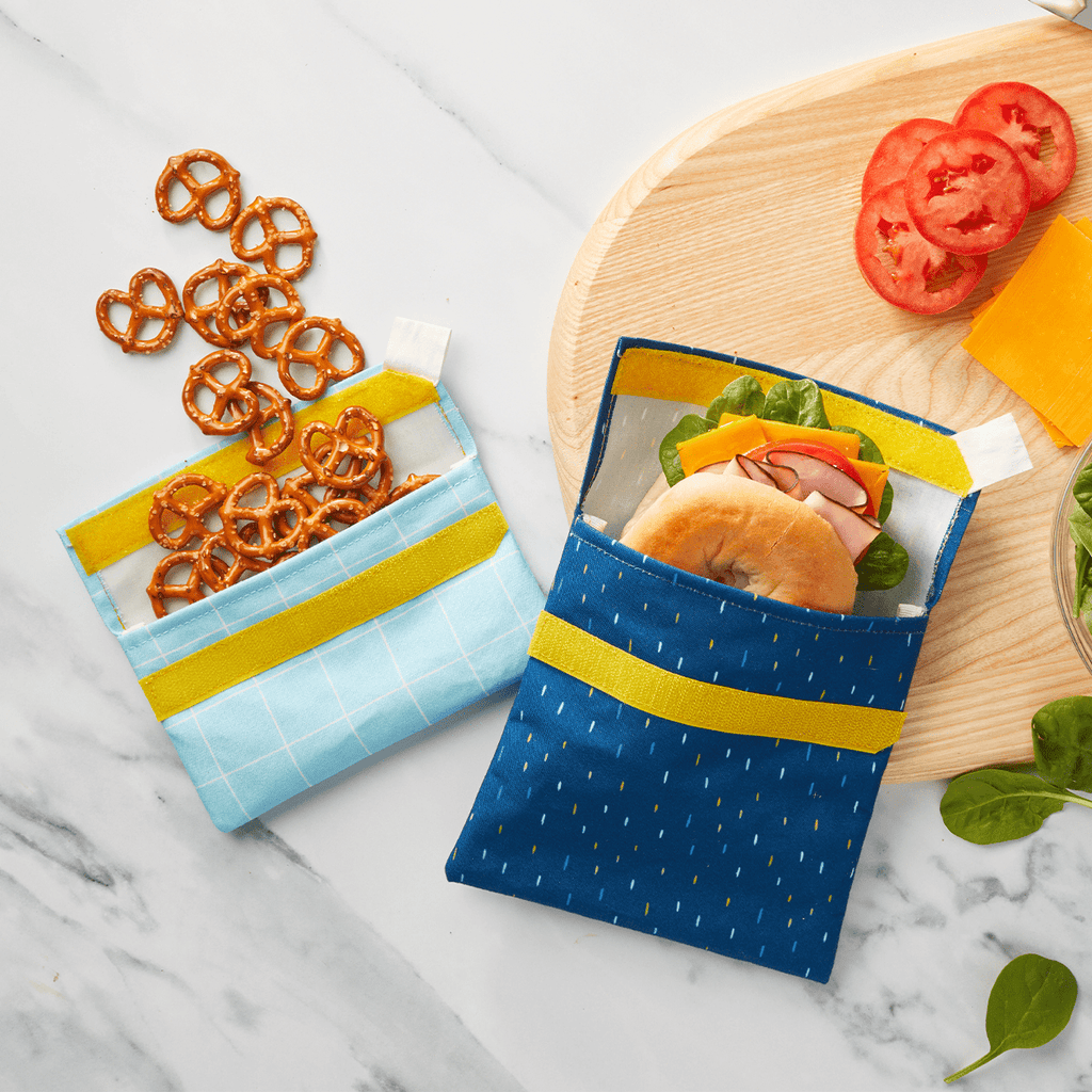 Snack packs in Ocean Dashes colorway opened with smaller one filled with pretzels spilling out, and larger one filled with a ham bagel sandwich. 